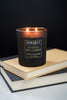 A black candle with a wooden lid, labeled 'Perfect Gentleman', burning with two lit wicks, placed on a stack of books.